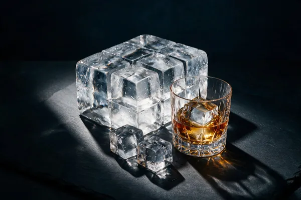 Stora kristallklara isbitar och ett rocksglas med whisky och en klar isbit på mörk skiffer