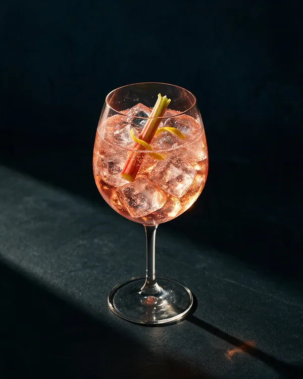 Ljusrosa Rabarber Spritz i vinglas med is, bubblor och en tunn rabarberstjälk